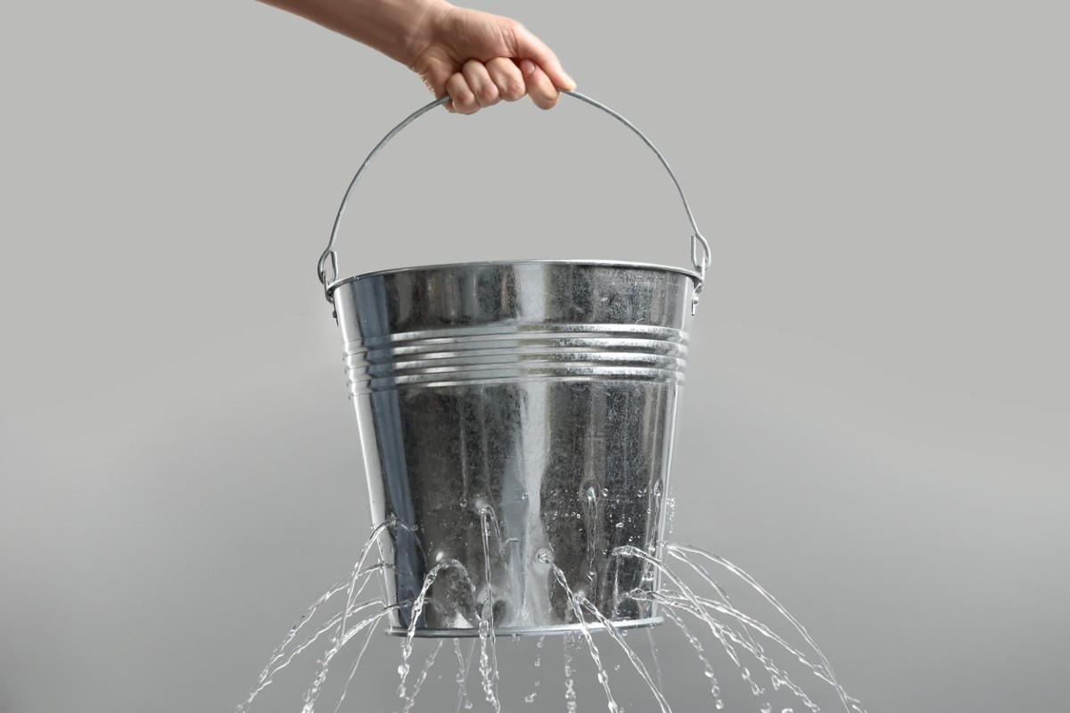 Woman holding leaky bucket with water on light grey background